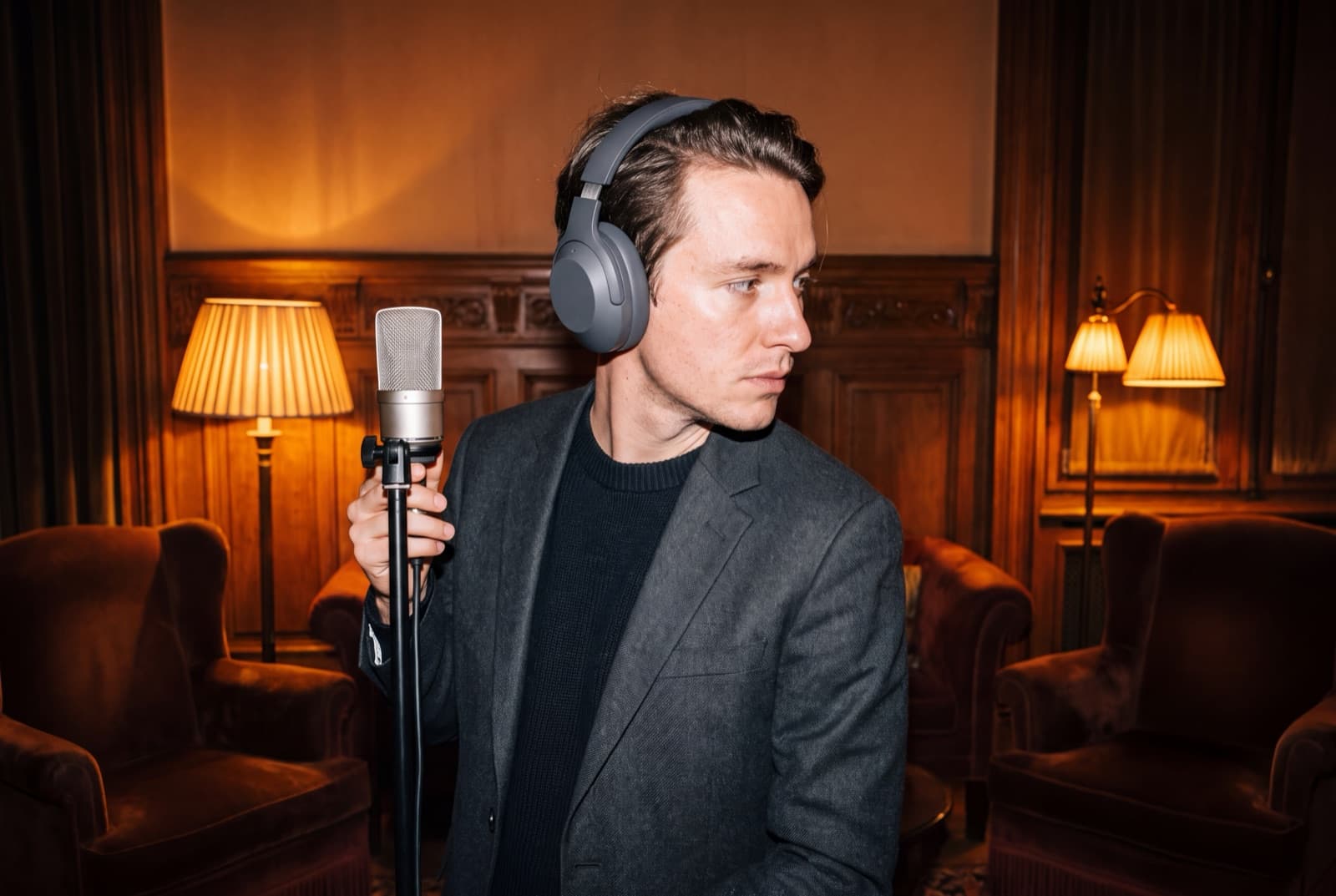 A media operator wearing clean headphones pauses beside a microphone in a warm wood-paneled room